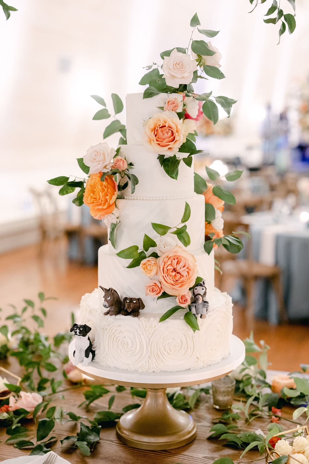A four-tier wedding cake adorned with cascading peach roses and custom pet figurines on the bottom tier