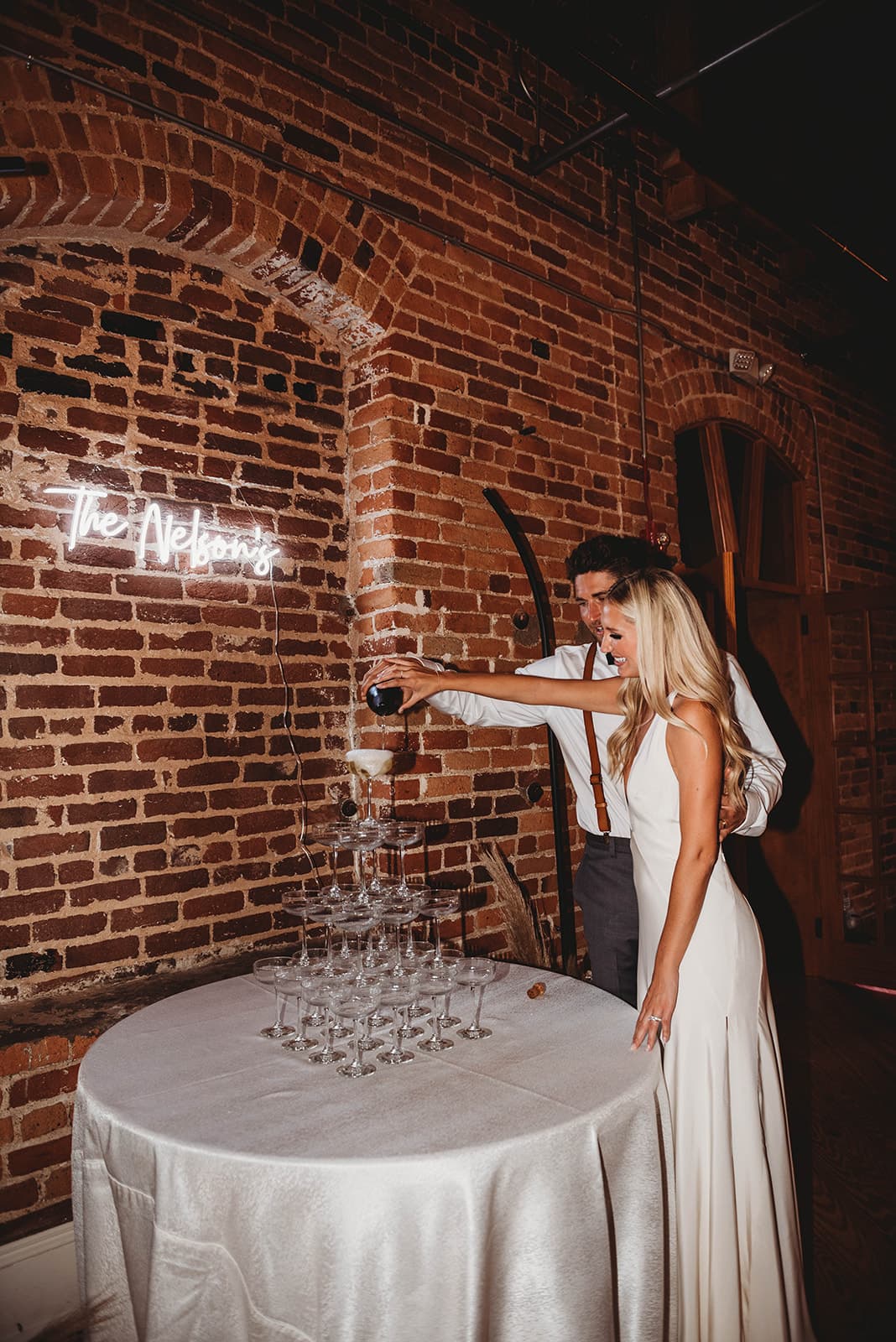 A couple pouring champagne into a tower of coupes at their wedding reception — Photo by Beasley Rustic Photography